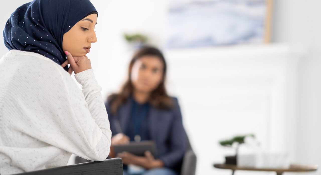A young Muslim woman sits in a chair with her head resting on her hand as she looks despondently across the room during a therapy session. She is dressed casually and is wearing a Hijab as she sits speechless in the session. Her female therapist is seated across from her as she tries to help her work through the struggles.